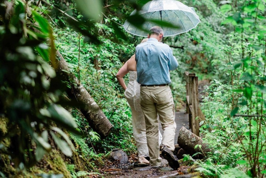 In a Pisgah National Forest Adventure Wedding near Lake Toxaway and the Greystone Inn, a groom holds a clear umbrella over the bride as they hike a trail in their wedding clothes. The bride lifts her dress to step over a puddle, and the trail is surrounded by bright green leaves and large felled trees.