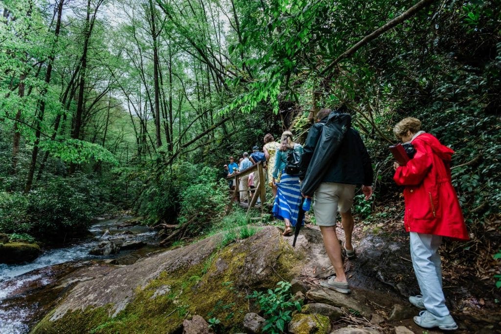 In a small outdoor wedding near Brevard, Lake Toxaway and Grey Stone Inn, a couple about to be married walks a narrow hiking trail along a cascading creek on the way to Courthouse Falls, lined with vibrant green trees and shrubs. Their family members walk closely behind them, over a small footbridge