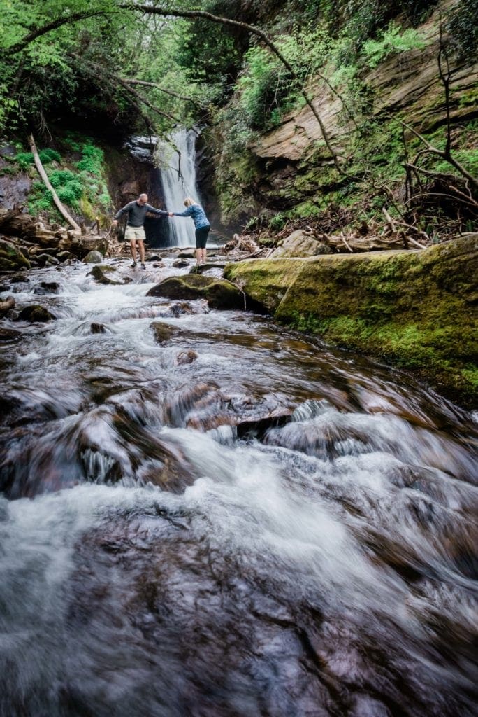 A couple explores a waterfall as a site for their elopement in Pisgah National Forest. Stream pours over mossy rocks, as the man offers a hand to the woman as she steps across the stream. A waterfall crashes directly behind them.