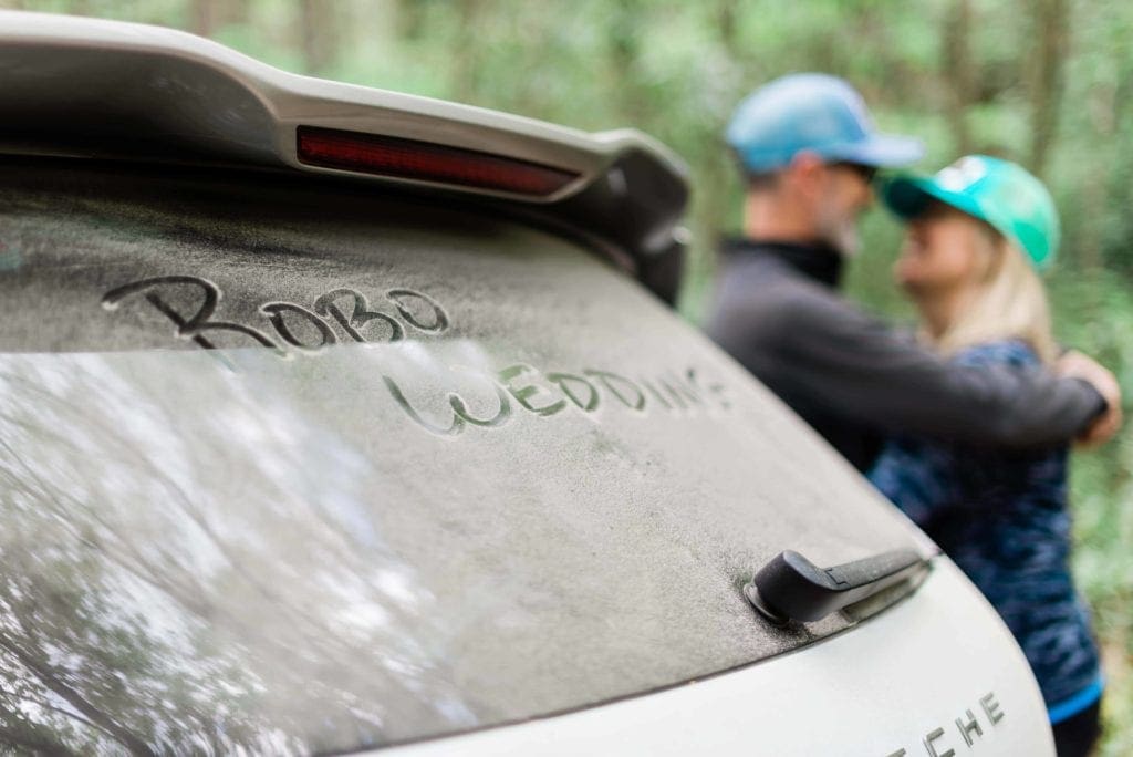 A couple snuggles in the background with trees behind them, while their car is dusty from driving on the forest service dirt roads has "RoBo Wedding" scrawled in the dirt on the back window