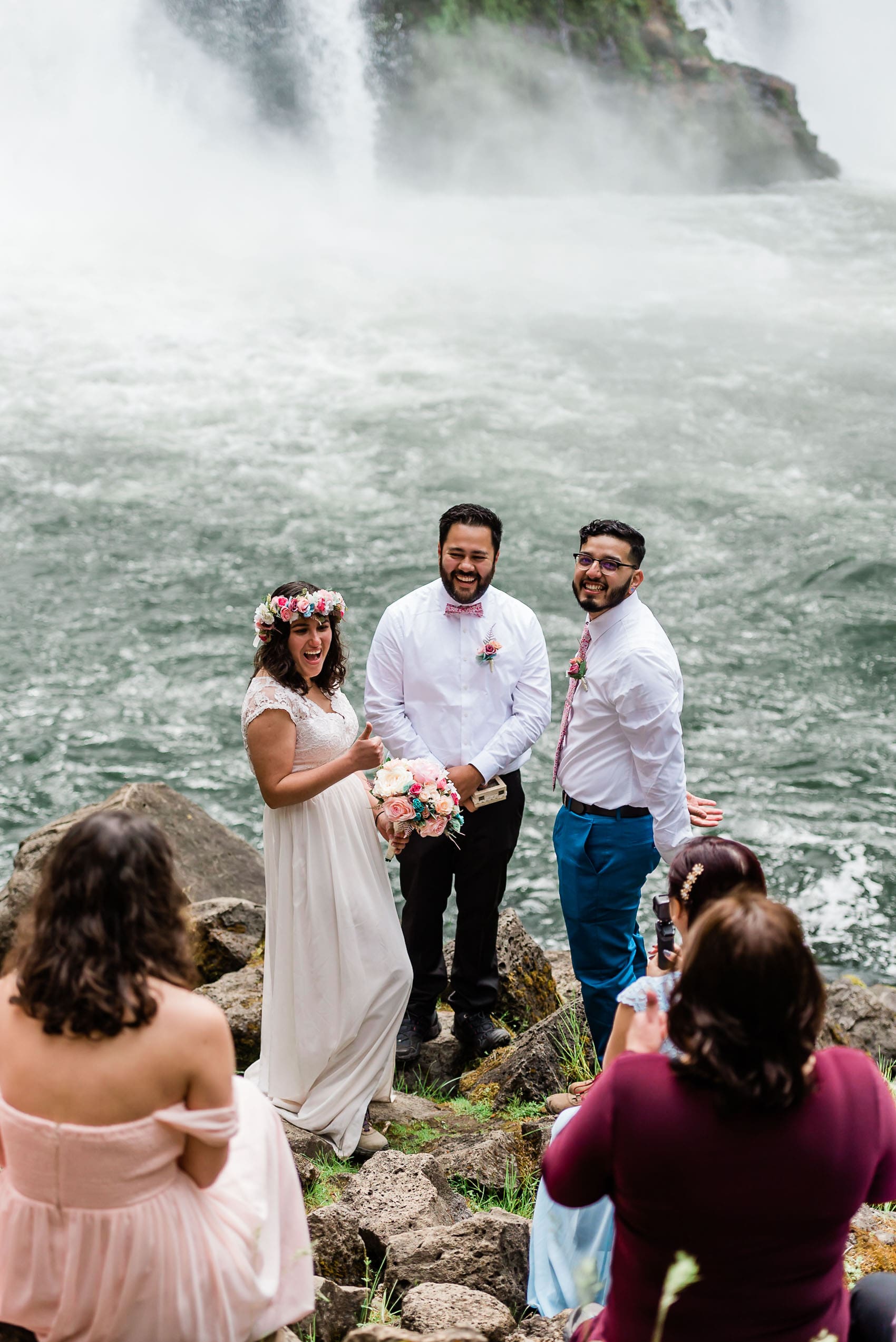 This picture is of an elopement experience. The bride and groom are standing at their alter, a rock in front of a large waterfall. They are standing with their officiant and we can see their small wedding party.