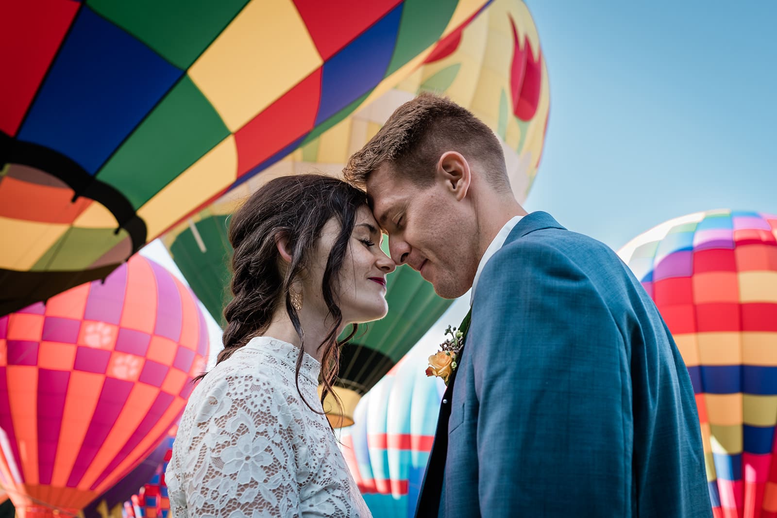 This is a picture of a bride and groom with their heads close to one another, touching foreheads. Behind them are a lot of hot air balloons.