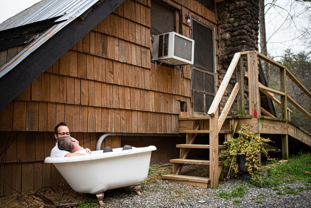 This is a picture of two men laying in a tub together outside. They are fully dressed and laughing together.