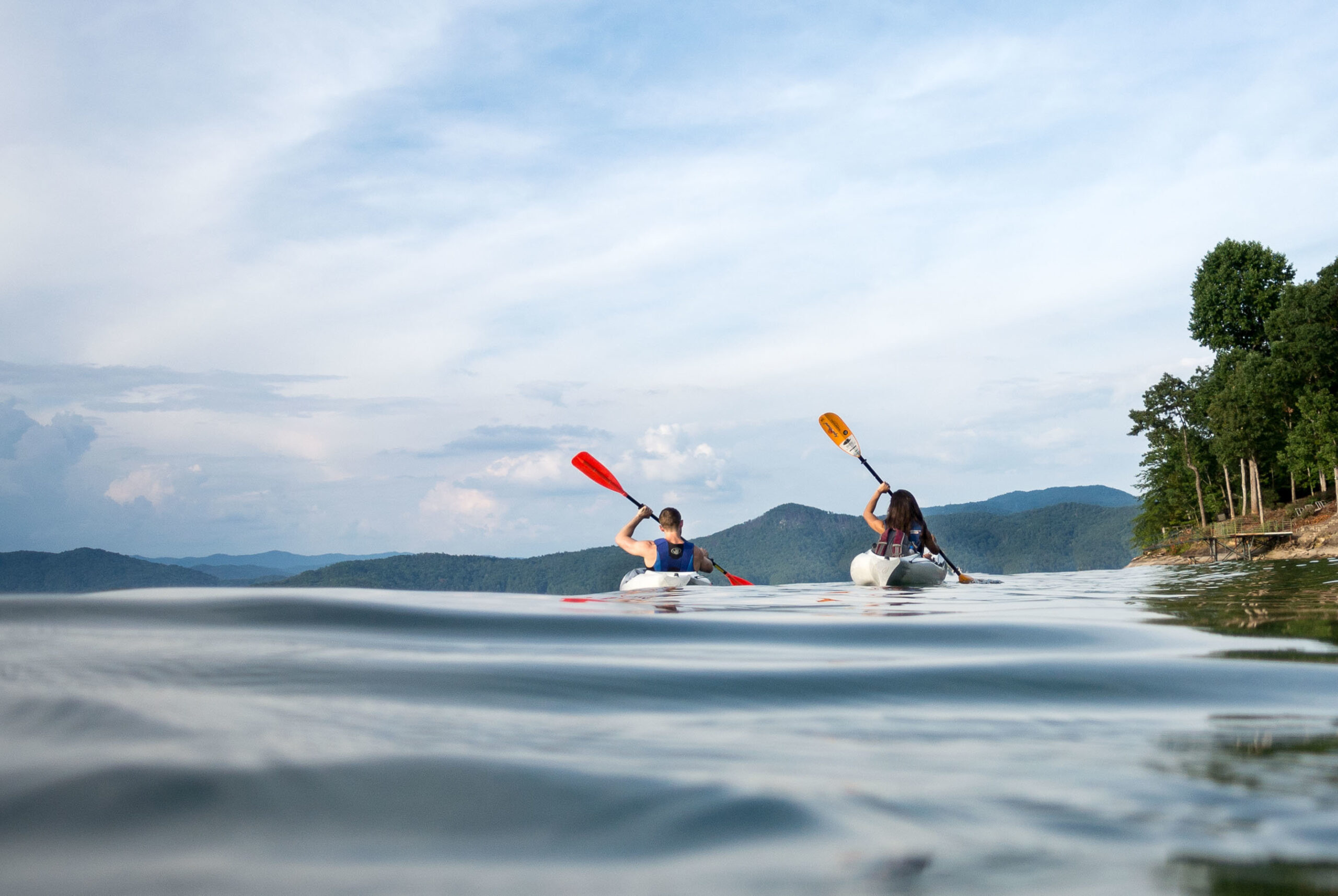 A bride and groom paddle kayaks along the shoreline the shimmering Lake Jocassee after their sunrise eloepment vows as captured by their adventurous luxury elopement photographer