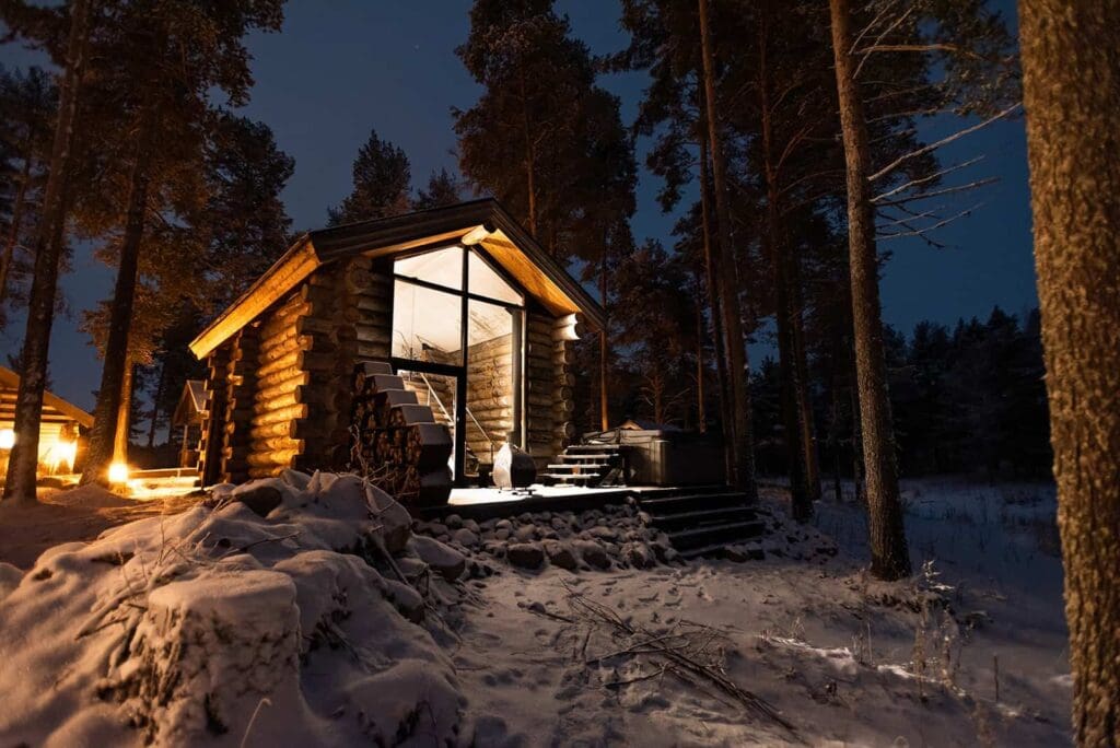 beautiful glass front cabin lit up at night, glowing warm light into the snow in an adventure elopement package for an elopement photographer who travels and can photograph the aurora borealis in alaska or sweden