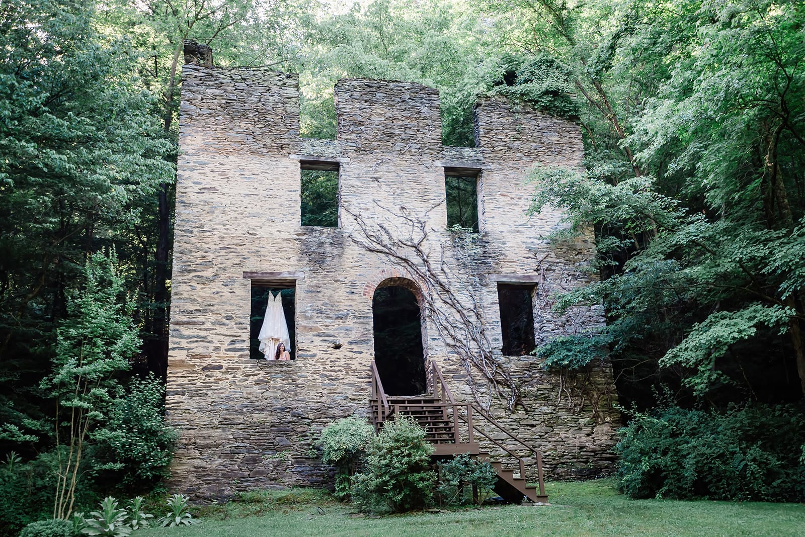 This is a picture of a wedding dress hanging from the window of an abandoned castle in the woods.