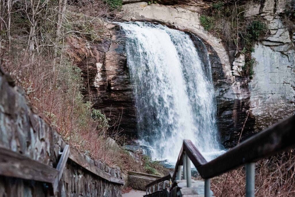 A picture looking down the wood and stone stairs to Looking Glass Falls, an elopement location off near the Blue Ridge Parkway.  The falls is rushing full, in a huge white cascade down the dark rock, tumbling into a boulder-filled pool at teh bottom.  Trees are  without their leaves in the early spring conditions.  This is a location where couples would need to plan how to adjust wedding plans after Hurricane Helene.