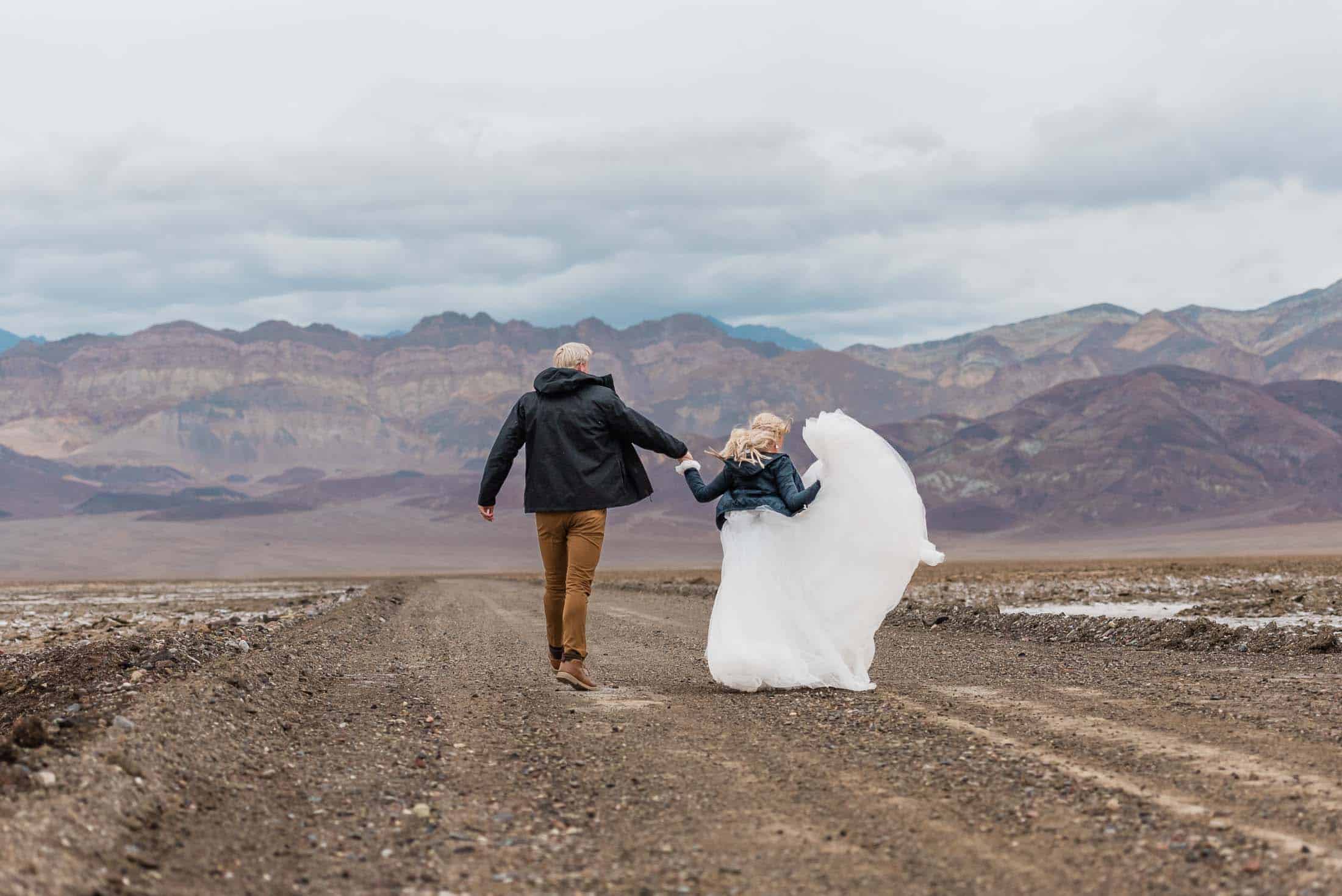 A groom and bride are running down a dirt road in the desert in Death Valley, away from the camera towards a mountain range edged by a salt pan. they are both in jackets as it has been raining, and the sky is dark and moody. the bride's train of her dress is flying freely around her as they run. It is a carefree scene