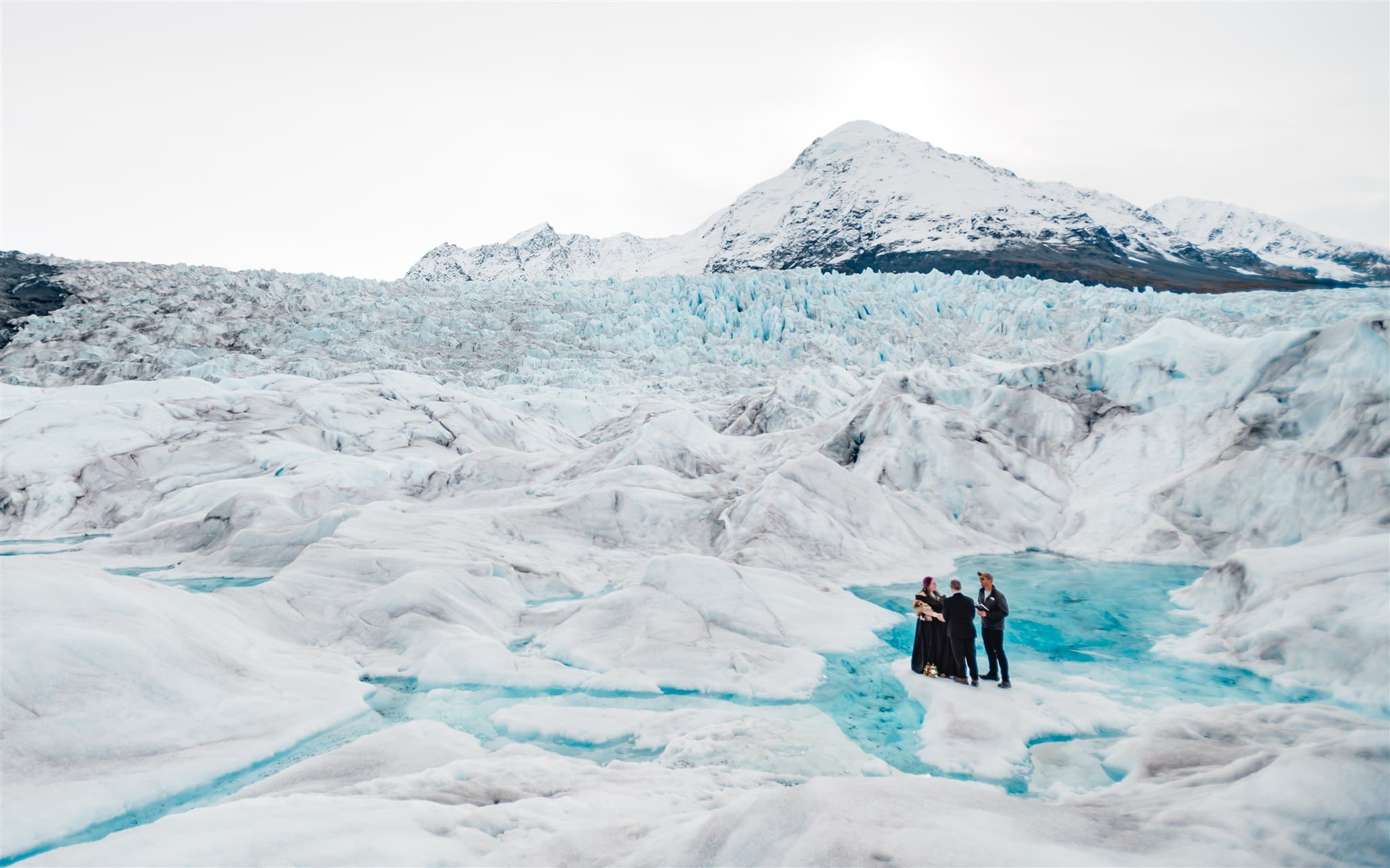 during a glacier elopement in alaska, a bride in a long black wedding dress, a groom in a black tux with a purple vest, and a wedding officiant holding a folio, wearing casual clothes are all standing on a white, icy glacier. there are jagged mountains and fields of icy spires behind them and around. next to them/behind them is a bright turquoise blue glacier melt "blue pool" .