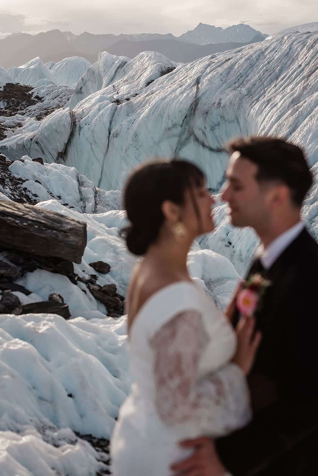 Softly focused, a bride and groom embrace in the foreground of the image in their formal wedding garments. Behind them, in focus, is an epic icy glacier with rolling mound of ice, cliffs of white, rocky veins, and blue tinted ice walls. The soft sunset lights the whole glacier wedding scene in a soft yellow light.