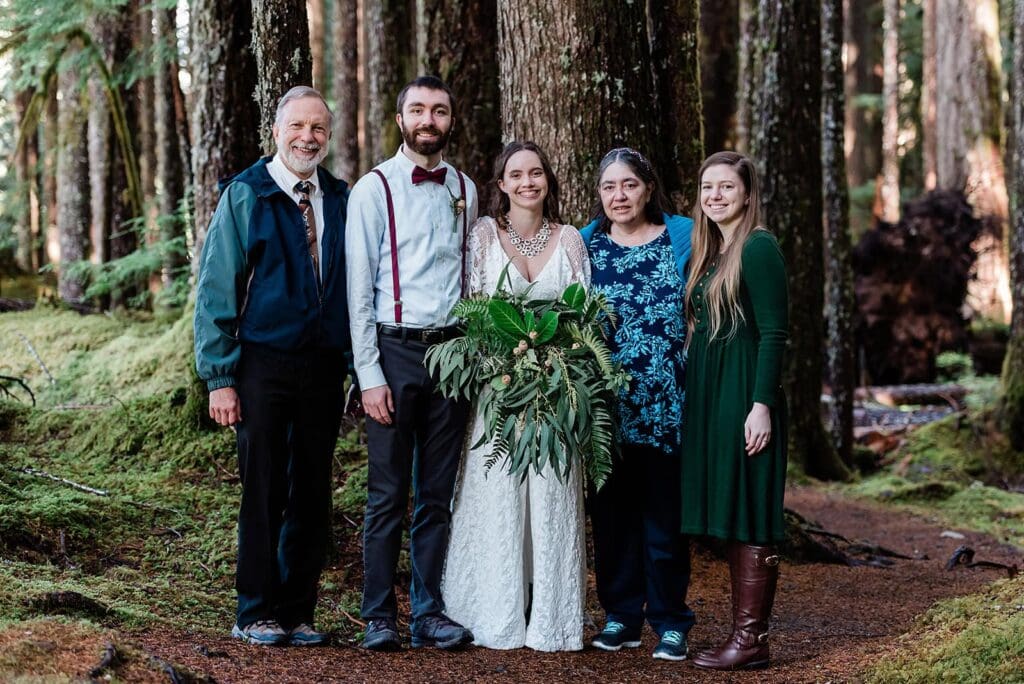 A family poses for a formal family photo in the Juneau Alaska type of lush green temperate rainforest.  There is moss and ferns all around them. 