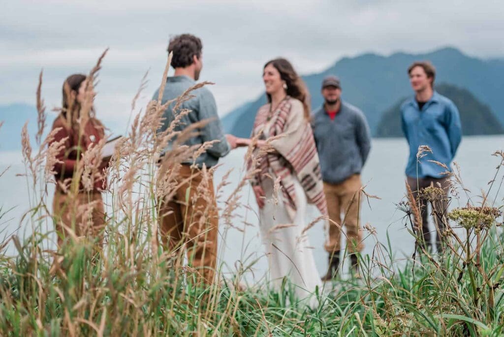 At an island in Resurrection Bay near Seward, Alaska, a couple says their vows with their few guests looking on with water behind them, and tall grasses flowing around and to the side of them. 