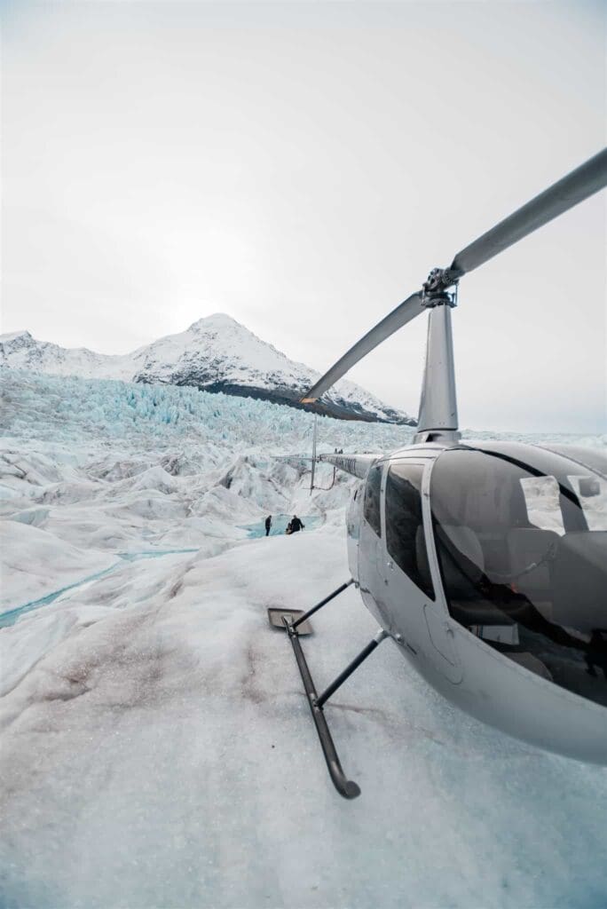 On the Knik Glacier, a white helicopter is in the front of the image frame from a luxury elopement in Alaska and behind it stretches a glacier in all directions. A tall snow covered peak rises up from the glacier in the background, and a ways behind the glacier you can see the couple with their officiant during the elopement ceremony on the glacier.