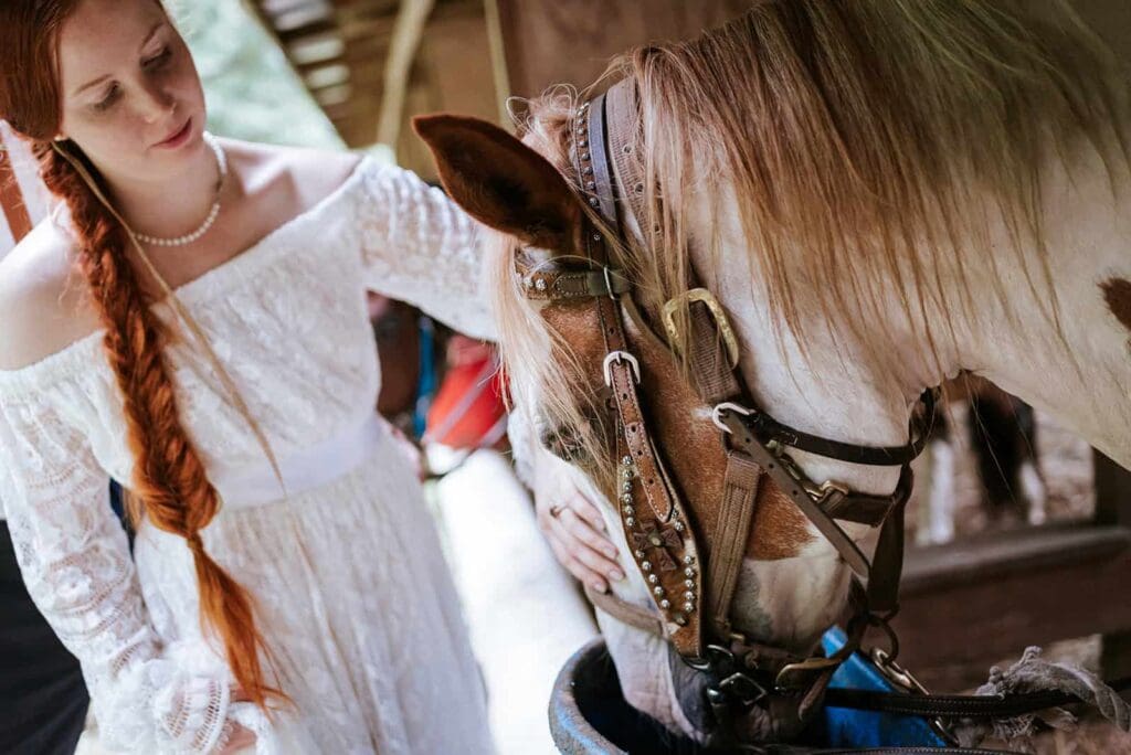 An eloping bride near Anchorage Alaska with long red hair in a boho style braid. She pets the head of a large horse that is drinking water back in the barn after their horseback ride for their adventure luxe elopement activity in Alaska.
