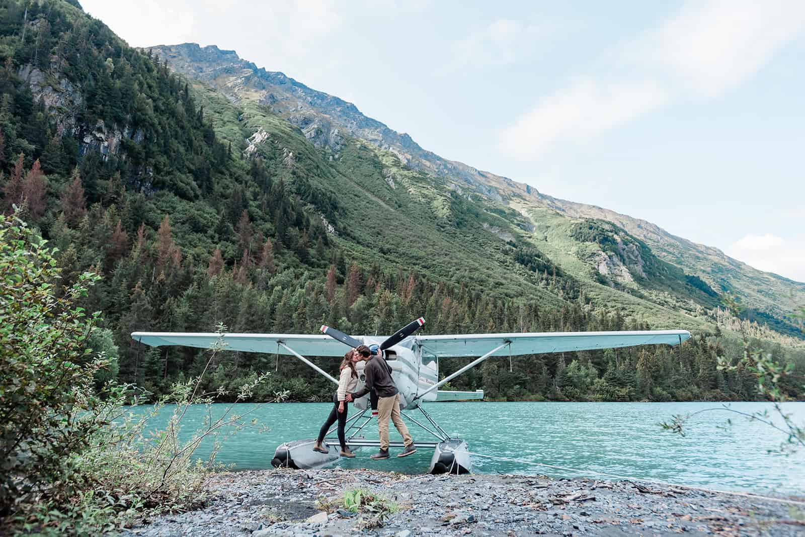 During a romantic vaction in Alaska, a woman and a man are each standing on a metal float skis of a large, white float plane that's pulled partially ashore, with part on the beach and part in the milky turquoise water of a high alpine lake. They are in casual clothes, and leaning to the middle from each side and kssing. The shoreline in front of them is small, gray pebbles and rocks. The mountain behind them is a rugged but lush green. The sky is light blue with wispy clouds.