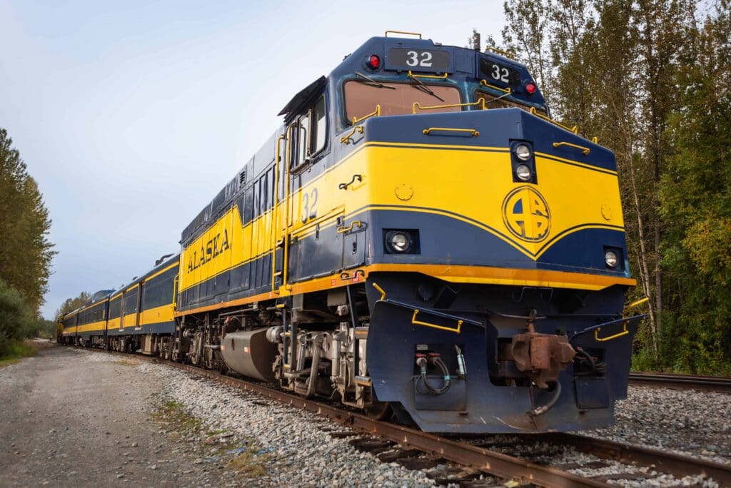 Before loading up for their train adventure luxe elopement in Talkeetna, Alaska, the front lead engine of a long blue and yellow Alaska Railroad train is visible, with a line of train cars stretching behind.