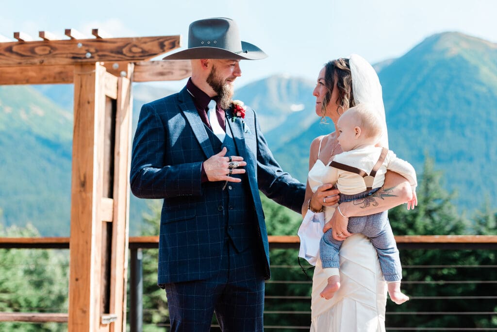 A couple holds their toddler while they elope in Alaska. They have just done their first look so each are in their formal wedding clothes, and faces are emotional. They Chugach mountains rise up behind them