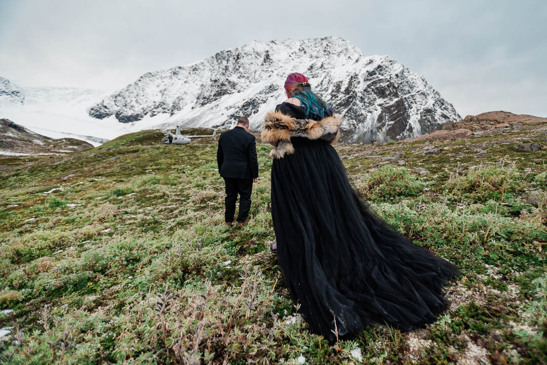 A bride and groom walk away from the photographer, and over a green and brown tundra of short grasses during their Alaska Elopement. She is wearing a black dress with a dramatic tulle train sweeping the earth, a fur stole, and has gorgeous rainbow colored long hair. The groom is wearing a black suit, and in the distance craggy snow covered Chugach mountains fill the frame. A tiny silver helicopter sits at the base of the mountain.
