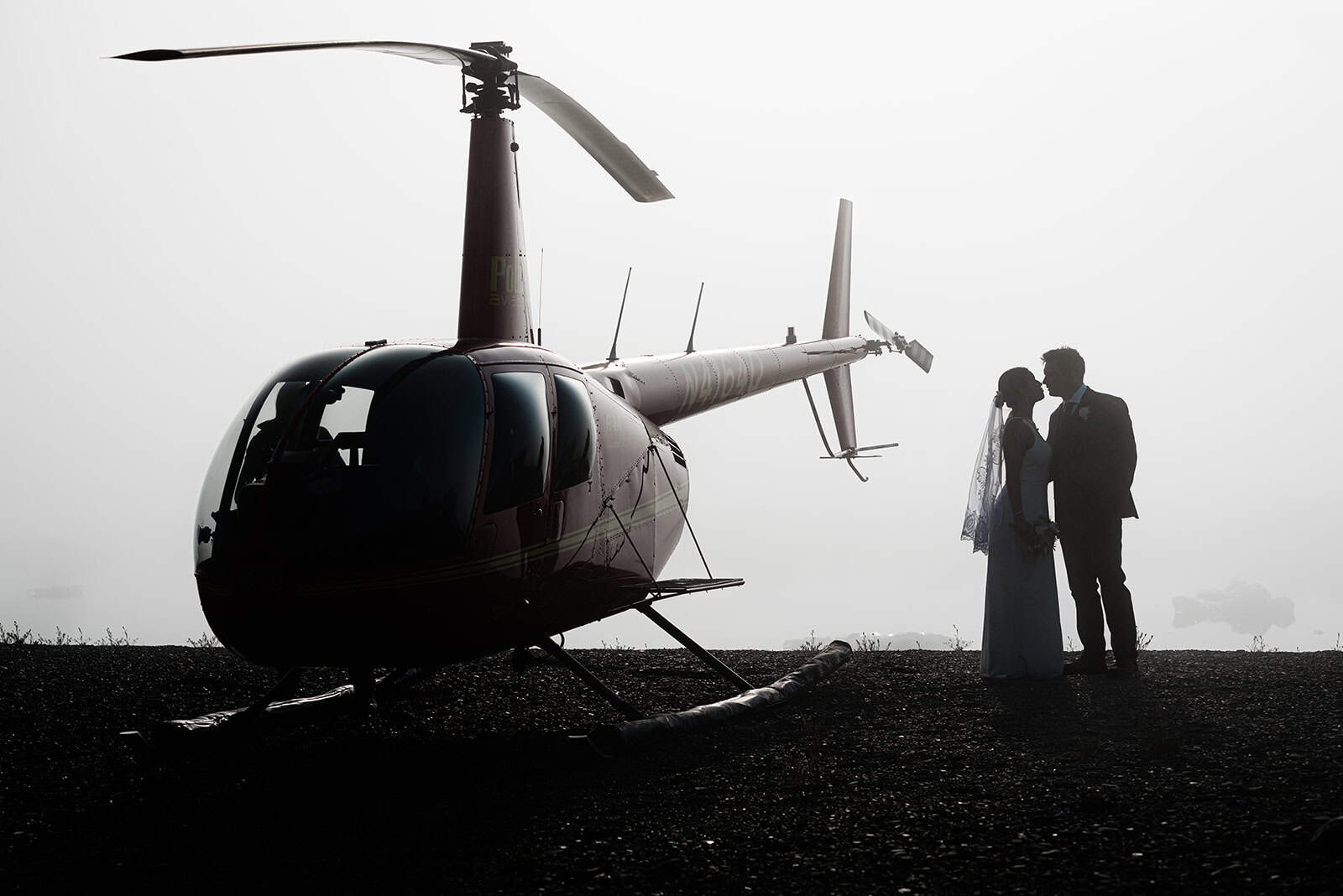A luxury private helicopter elopement flies over Bear Glacier lands on the shore of the iceberg lagoon near Seward Alaska. A bride and groom in wedding formal wear are standing next to the red helicopter, but everything (including an iceberg in the water behind them) is shiloutted in a fog so it feels eerie, romantic, and looks almost black and white.