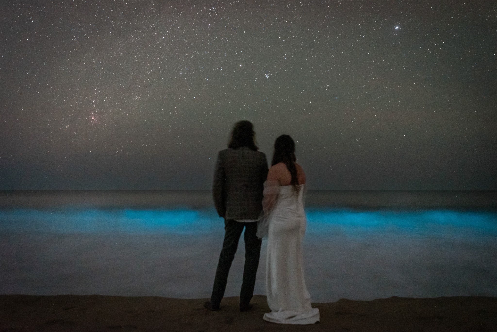 A bride and groom in their wedding finest stand in the dark night during their adventurous luxury Oaxaca Mexico Elopement. The night sky is littered with stars and in front of them filling the bottom half of the frame, a bright blue and milky white bioluminescent wave crashes ashore.