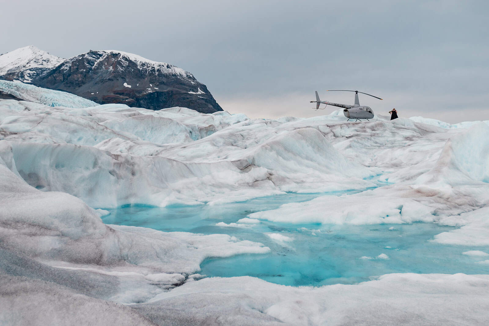 A bride in a black dress walks over the white ice during a Knik Glacier elopement towards her helicopter. the front of the frame is filled by a bright blue glacier melt pool, and dark craggy mountains hulk in the background beyond the heli