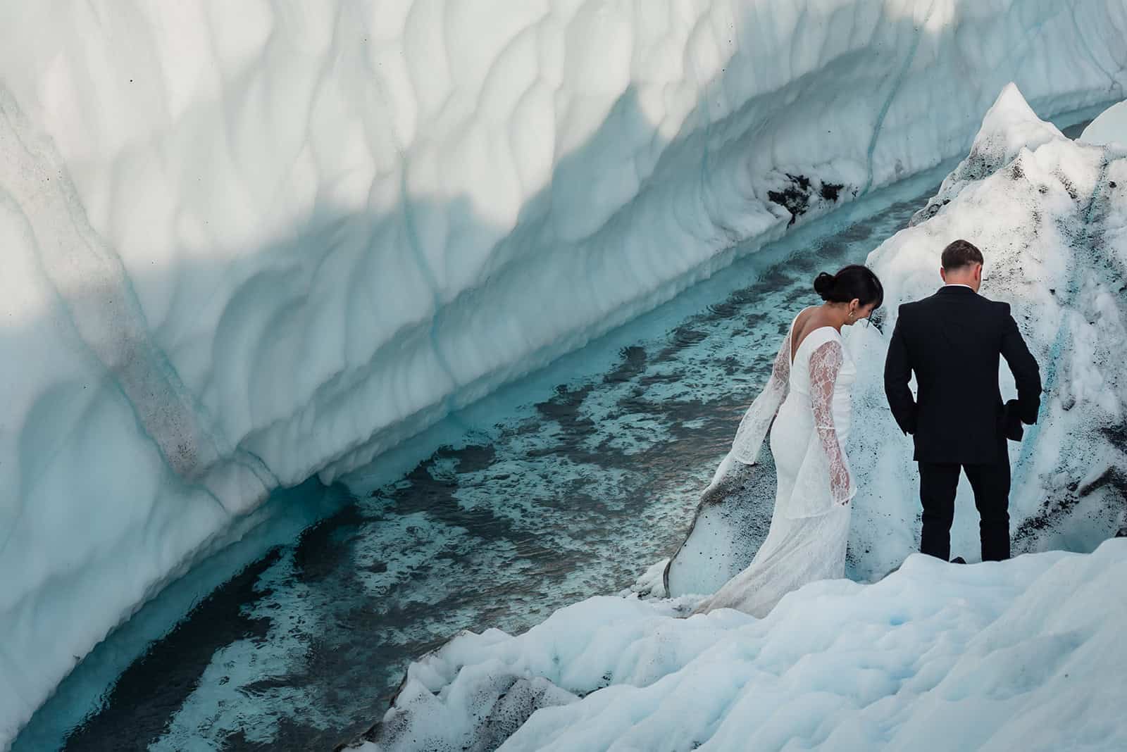 The bride and groom in their formal wear walk on the icy white Matanuska Glacier with the midnight sun casting shadows of icy ridges against ice cliffs that fill the frame behind them. below them. A bright blue glacier meltwater stream flows towards where they will exchange their vows while eloping on the glacier.