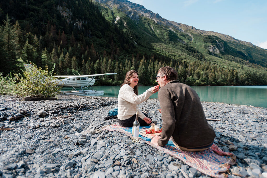 A couple enjoys a fun picnic on a rocky beach of an lake in the mountains during their summer multi-day elopement in Alaska.  The float plane they traveled on is visible in the distance behind them as they sit and laugh on the shore. 