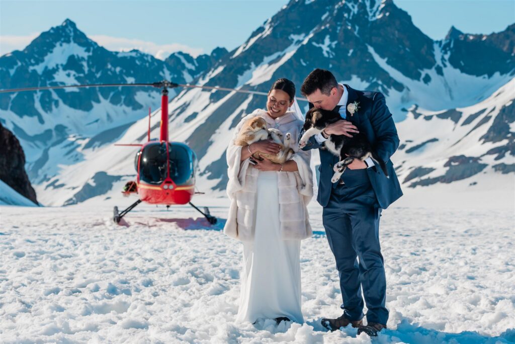 A bride and groom in are in their wedding formals, holding sled dog puppies during their summer dogsledding elopement on a glacier in Alaska.  A helicopter took them to the Godwin glacier with bright white snow around them, dark jagged mountains behind them, and the bright red helicopter visible just off the bride's left shoulder.