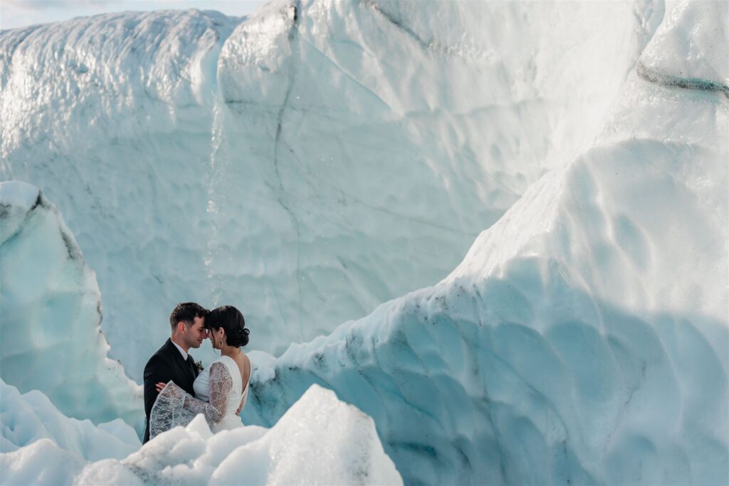 This is a picture of a bride and groom having an intimate moment tucked in the white and blue glacier ice walls in the beauty of the Matanuska glacier. Behind, you can see the melting ice streaming down. The texture of the glacier is mesmerizing.