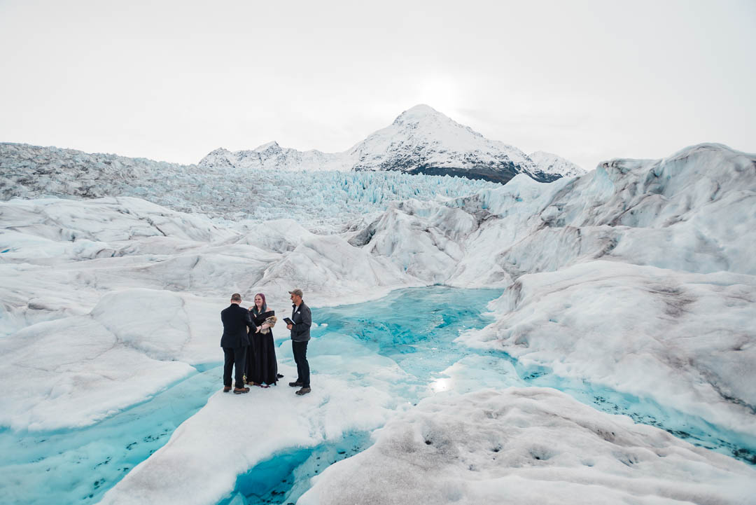 A couple and their officiant stand on the white ice surrounded by a bright blue glacier blue pool on the Knik Glacier during their Alaska glacier elopement. There are jagged spires of glacier ice on one side of them, bright blue water on three sides, and a tall, snow covered mountain behind them as they read their vows.