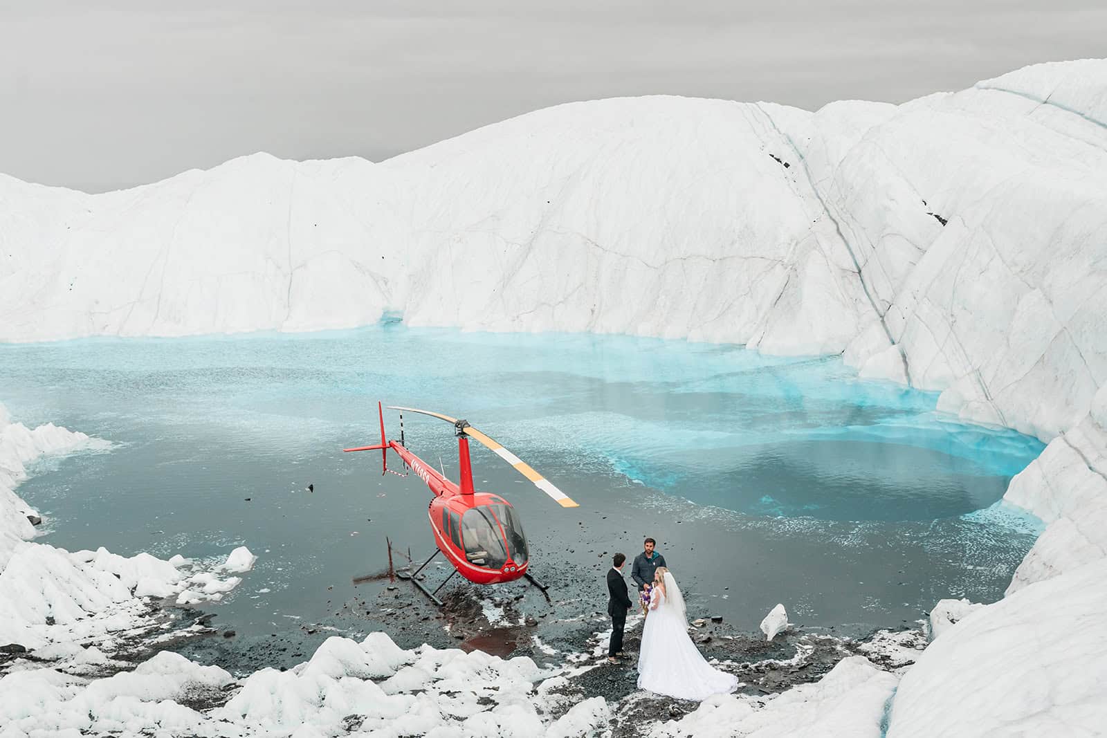 Wedding couple celebrating on Matanuska Glacier standing on a icy white glacier next to a bright blue glacier blue pool next to their red helicopter.