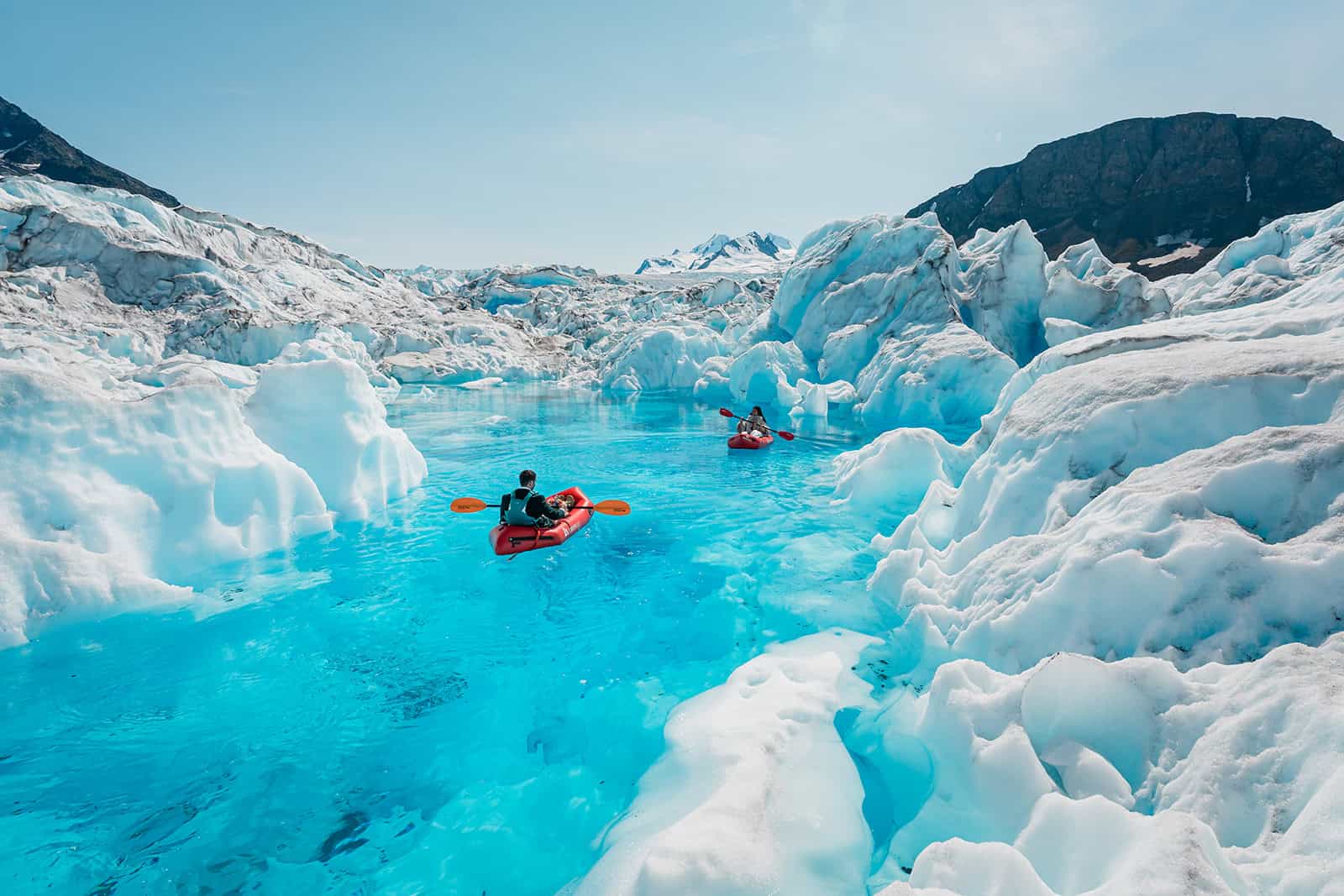 an eloping couple is kayaking in red packrafts in a bright blue "glacial melt blue pool" on the Knik Glacier. Surrounding the bluepool is jagged glacier ice with white and blue peaks of ice.