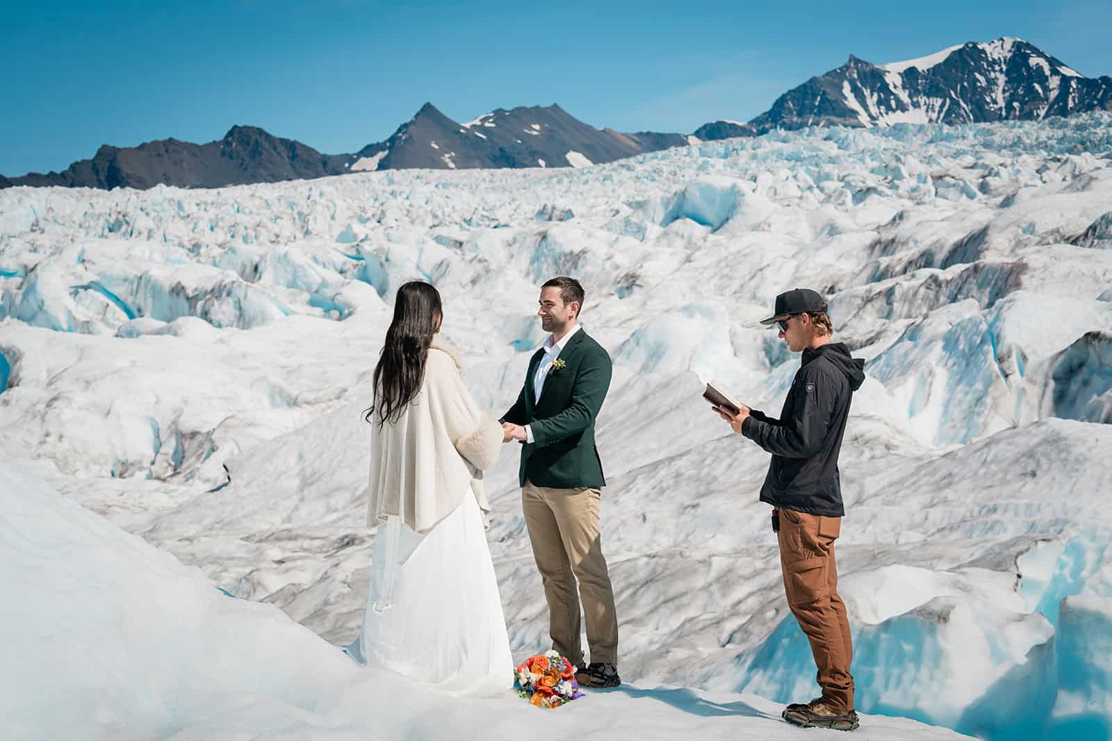 an eloping couple exchange vows with their helicopter pilot as their officiant standing atop the Knik Glacier. a jagged icefall is behind them with white peaks of ice, and below them to their right is the bright blue water of the Knik Glacier's famous bluepools.